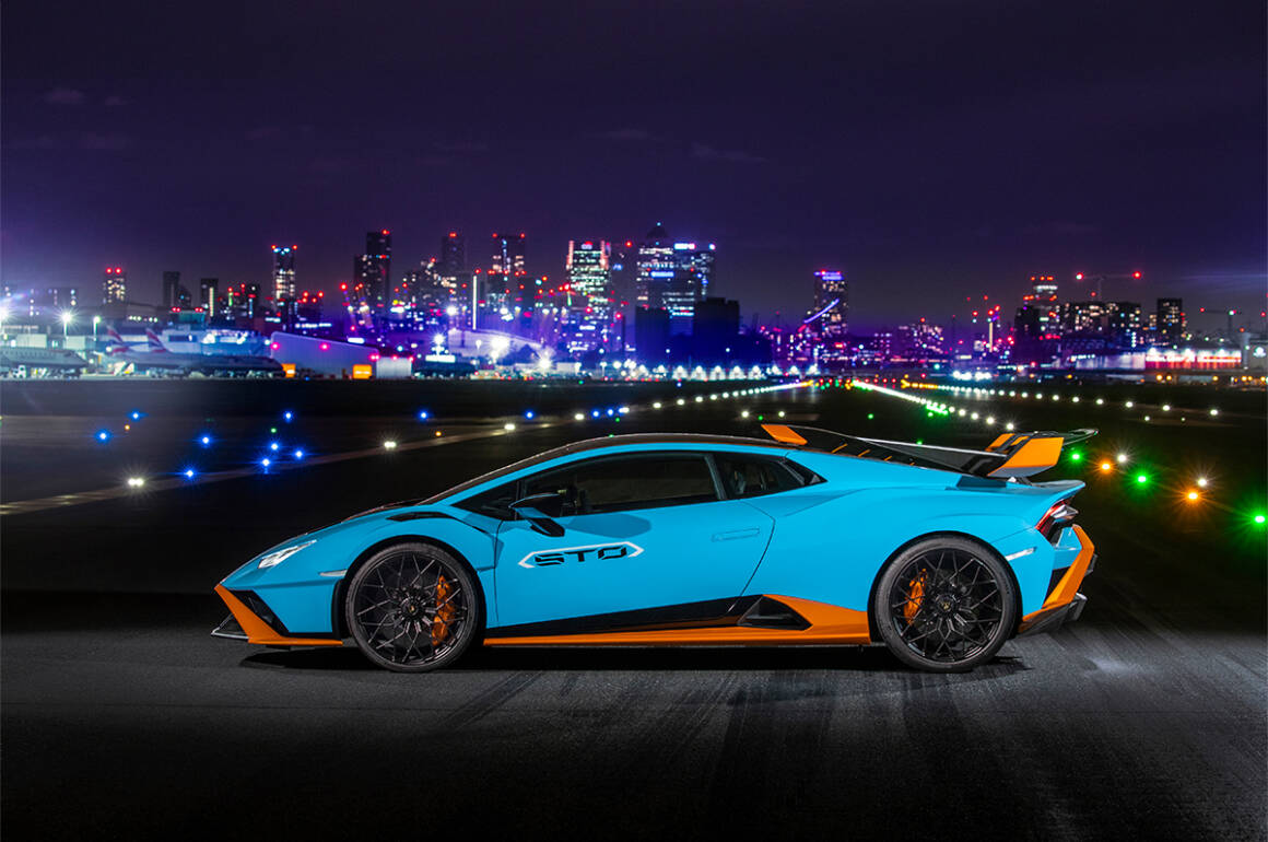 A blue and orange Lamborghini on a road at night with a lit up skyline behind it