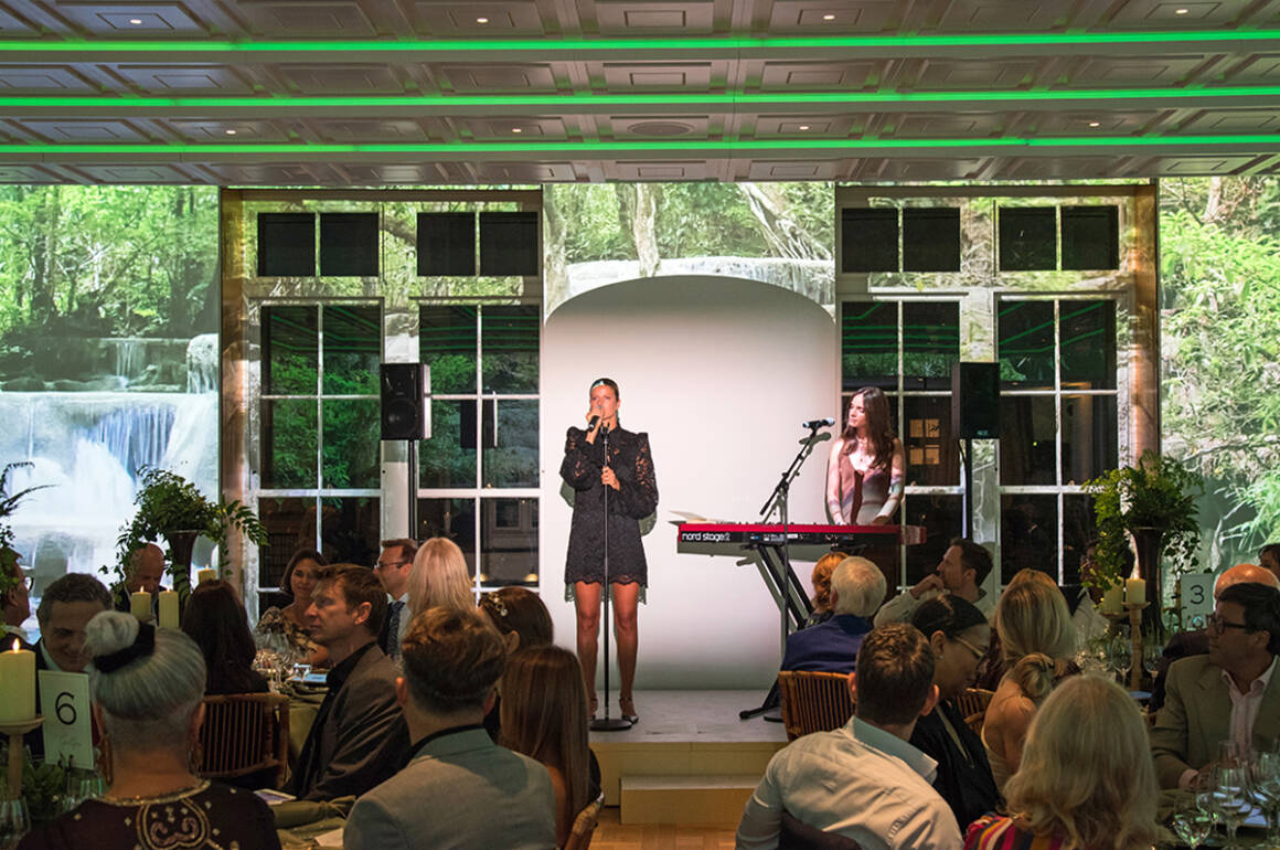 A woman singing on stage next to a person on a keyboard with green lights above her and people watching sitting at tables