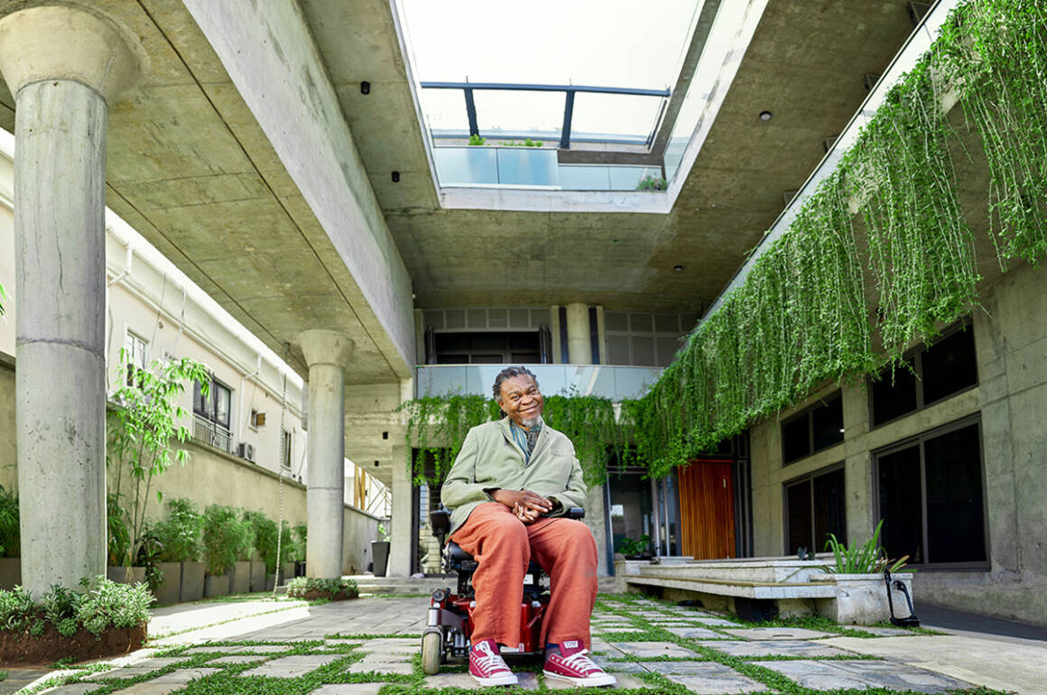 A man sitting in a wheelchair, wearing red trousers, in a courtyard of a building with plants hanging over the interior balconies
